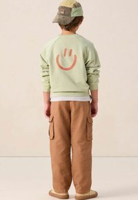Kind met een groene sweatshirt met een oranje glimlachend gezicht op de achterkant, bruine cargo-broek, een patchwork pet en beige sneakers, staande met de rug naar ons toe.