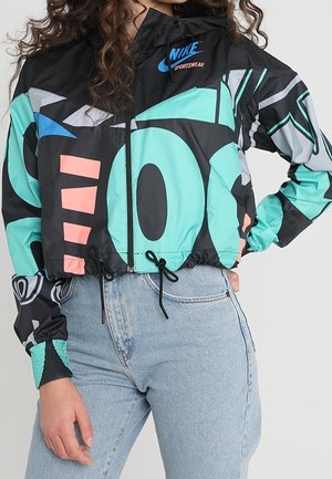 Femme portant une veste de sport noire Nike avec des motifs abstraits colorés et un jean bleu clair, mains sur les hanches.