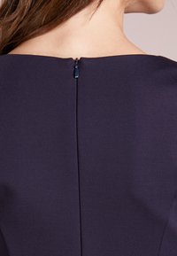 Mörkblå klänning med hög halsringning och en dold dragkedja i ryggen. Slät tygtextur med minimal sömnadsdetalj.