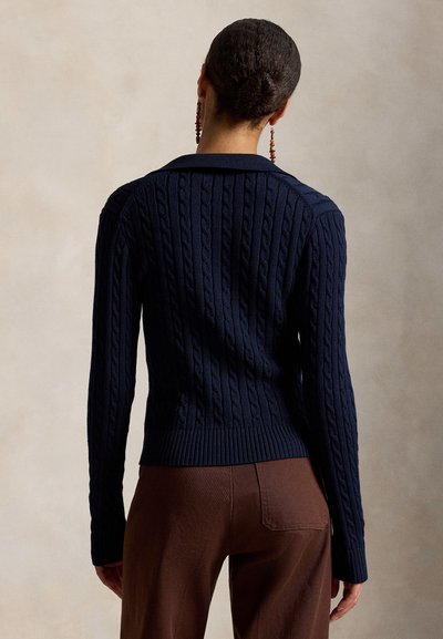 Sötét hajú nő alacsony kontyban, tengerészkék fonott mintás pulóvert és barna nadrágot visel, háttal látható, semleges háttér előtt.