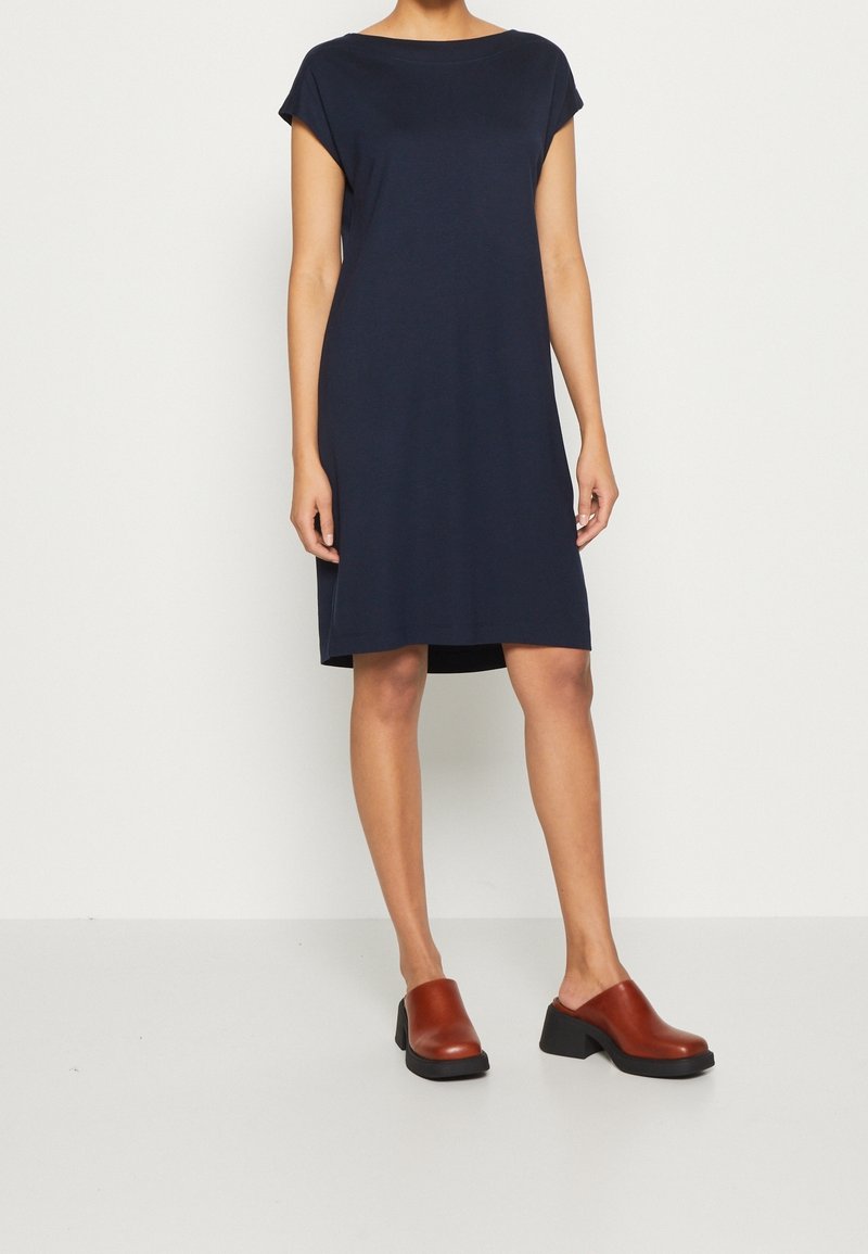 Femme portant une robe bleue marine simple longueur genou avec des manches courtes et des chaussures marron à talons sans lacets, debout contre un mur uni.