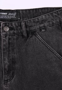 Cintura de pantalones vaqueros de mezclilla negra con botón de metal, trabilla para cinturón, bolsillo delantero y etiqueta que dice "FORMER" y talla 32.