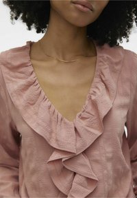 Haut rose avec un profond décolleté en V orné d'un volant. Le tissu présente un motif texturé qui ajoute de la dimension. Collier simple et délicat en or exposé.