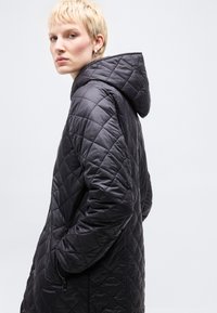 Γυναίκα με κοντά ξανθά μαλλιά που φοράει μαύρο καπιτονέ μπουφάν με κουκούλα, γυρισμένη στο πλάι με τα χέρια στις τσέπες, μπροστά σε απλό ανοιχτό φόντο.