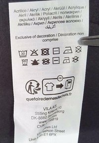 Tag med tvättråd, som visar vårdsymboler, material på flera språk och företagsinformation. Vit tygbakgrund.