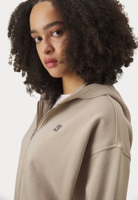 Haut zippé beige en tissu texturé, doté d'une capuche et d'un petit logo sur la poitrine. Le design comprend une coupe décontractée et des accents côtelés.