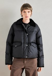 Črni napihnjen plašč s ovratnikom, sprednjimi gumbi, stranskimi žepi in sijočim zaključkom. Nošen je preko črne puloverja in rjavih hlač.