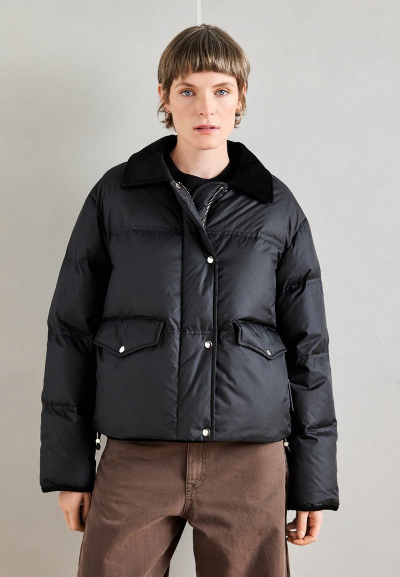 Črni napihnjen plašč s ovratnikom, sprednjimi gumbi, stranskimi žepi in sijočim zaključkom. Nošen je preko črne puloverja in rjavih hlač.