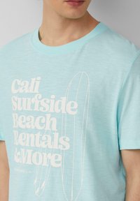 Lichtblauw katoenen t-shirt met korte mouwen, voorzien van witte grafische tekst en surfplankillustraties op de voorkant.