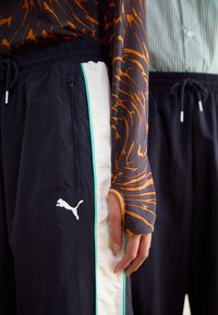 Μαύρο αθλητικό παντελόνι με λευκές και τιρκουάζ πλευρικές πάνελ, διαθέτει τσέπη με φερμουάρ και λογότυπο Puma. Συμπληρώνεται από ένα μοτίβο μακρυμάνικο τοπ.