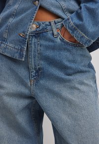 Džinsinė striukė su šviesesniu mėlynu atspalviu, turinti sidabrinius aksesuarus ir oranžinius siūlus. Džinsai turi laisvą kirpimą ir klasikinį penkių kišenių dizainą.