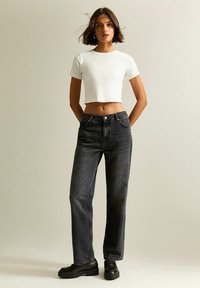 Mujer de pie con las manos detrás de la espalda, vestida con una camiseta corta blanca, jeans negros de talle alto y mocasines negros sobre un fondo plano.