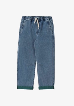 Enfant aux cheveux bouclés tirant un sweat-shirt vert par-dessus la tête, portant un jean bleu ample et des baskets beige sur un fond beige uni.