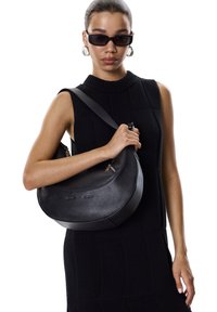 Sac en cuir noir porté à l'épaule avec une forme courbée en demi-lune ; il possède une texture lisse et une longue bandoulière. Le mannequin porte une robe noire sans manches.