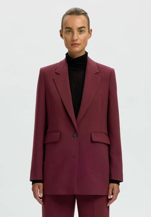 Vrouw met naar achteren gestyled haar, gekleed in een bordeauxrood colbert en pantalon over een zwarte coltrui, staand tegen een effen lichte achtergrond.