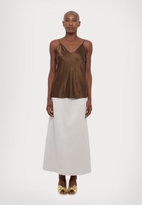 Brun sateng camisole-top med tynne stropper, kombinert med et langt hvitt skjørt og gule knyttesandaler, stående mot en enkel bakgrunn.