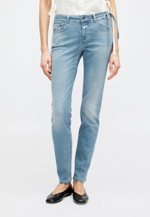 Jeans slim fit azzurri chiari indossati con ballerine nere, abbinati a un top bianco senza maniche con laccetti laterali.