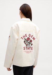 Cremefarvet jakke med en afslappet pasform. Har rød blomsterbroderi og tekst, der læser "THE GEM STATE" på ryggen. Bomuldsstof.