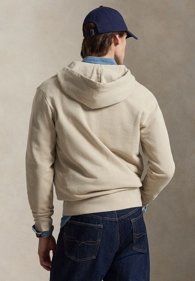 Man wearing beige hoodie, navy blue cap, denim shirt, and dark blue jeans facing away against neutral background.