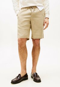Beige Shorts mit einem elastischen Bund und Kordelzug, die einen entspannten Schnitt und eine glatte Textur aufweisen. Kombiniert mit dunkelbraunen Slippern.