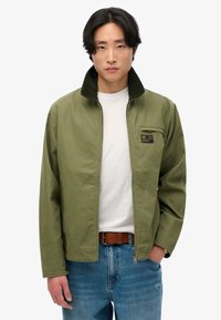 Olijfgroene jas met een corduroy kraag, voorrits en een kleine borstzak. Draagt over een wit shirt en gecombineerd met lichtblauwe jeans.