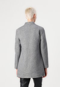 Abrigo gris con un diseño entallado, mangas largas y un cuello alto. Hecho de tela texturizada. Cae por debajo de las caderas. Líneas simples y limpias.