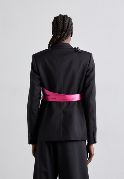 Blazer noir ajusté avec une ceinture en ruban rose. Le costume présente un design structuré et un détail de bouton unique à l'épaule.