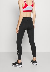 Leggings negros con una cinturilla alta y líneas de acento blancas en la parte inferior de las piernas. Combinados con un sujetador deportivo rojo y gris.