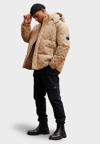 Béžová vzorovaná prošívaná bunda s kapucí, černé cargo kalhoty a černé šněrovací boty. Bunda má na rukávu nenápadné logo.