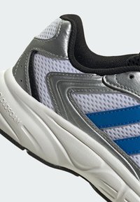 Gros plan d’une chaussure de sport blanche avec des rayures bleues, des surcouches argentées, une doublure intérieure noire et une semelle épaisse blanche rembourrée étiquetée « Cloudfoam ».