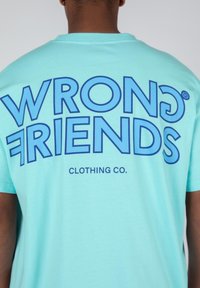 Camiseta de color azul claro con un gráfico en negrita que dice "AMIGOS INCORRECTOS" en azul oscuro, con "CLOTHING CO." en una tipografía más pequeña debajo. Textura de tela suave.