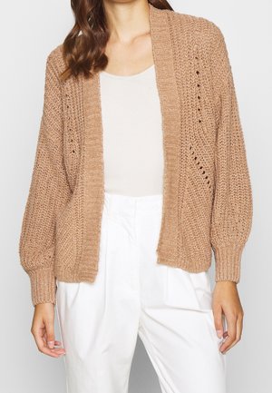 Femme portant un cardigan beige ouvert devant, tricoté, sur un top blanc sans manches et un pantalon blanc taille haute.
