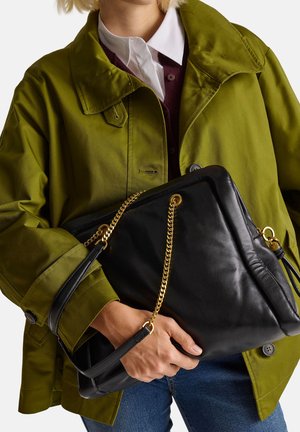 Schwarze Lederhandtasche mit einer goldenen Kettenriemen, gehalten von einer Person, die eine grüne Jacke und ein weißes Hemd trägt, mit sichtbarem Denim darunter.