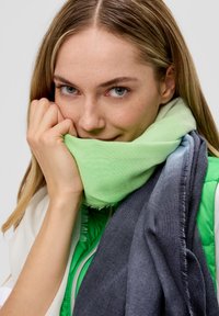 Lichtgroene katoenen sjaal gelaagd over een grijze sjaal, met franjes; model houdt de sjaal dicht bij haar gezicht en draagt een groene jas eronder.