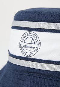 Cappello a secchiello blu navy con una fascia bianca e accenti argentati, dotato di un logo rotondo con testo e un design distintivo.