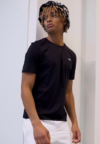 Camiseta negra de manga corta hecha de tela suave, con un logo rojo en el pecho. Combinada con pantalones blancos y un sombrero de cubo con estampado.