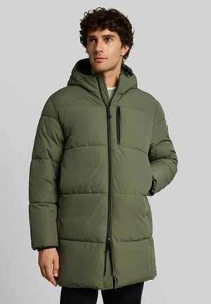 Homme portant une longue veste matelassée à capuche vert olive avec une fermeture éclair noire et la main dans la poche, regardant légèrement sur le côté.