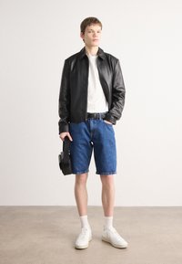 Svart skinnjakke, hvit t-skjorte, blå denimshorts, holder en svart bag, hvite sneakers med teksturert såle, ensfarget bakgrunn.