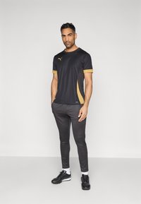 Μαύρο αθλητικό t-shirt με χρυσές λεπτομέρειες, κοντά μανίκια και στενή εφαρμογή, συνδυασμένο με γκρι παντελόνι jogger και μαύρα αθλητικά παπούτσια.