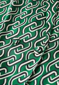 Grüner Stoff mit einem geometrischen Kettenmuster in Weiß und Marineblau, versehen mit zwei passenden, stoffbedeckten Knöpfen auf einer glatten Textur.