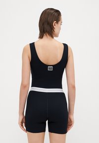 Črn rebrast bodysuit z izrezom na hrbtu, bel elastičen pas in oznaka z logotipom na zadnjem delu. Kratke tesno prilegajoče noge. Gladka tekstura.