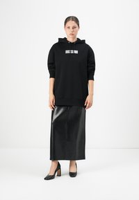 Žena koja nosi crnu oversized hoodie sa špagama, crnu kožnu midijevsku suknju i crne cipele s visokom petom, stoji naslonjena na običnu bijelu pozadinu.