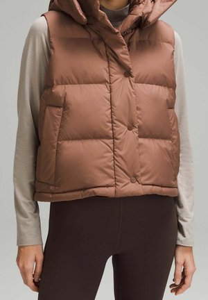 Femme portant un gilet matelassé sans manches marron avec des boutons-pression sur un haut beige à manches longues et des leggings marron foncé, debout devant un fond uni.