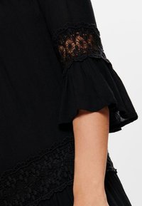 Vestido de tela negra con mangas acampanadas, que presenta detalles de encaje alrededor de los puños y el dobladillo, añadiendo textura y un patrón floral.
