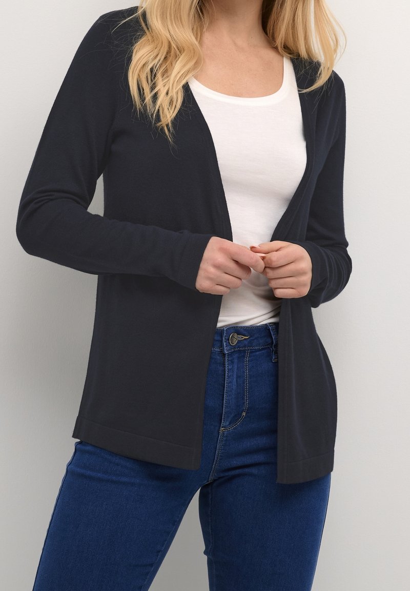 Femme portant un cardigan noir ouvert sur une chemise blanche et un jean bleu, les mains jointes près de la taille.