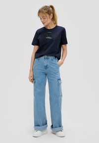 T-shirt bleu marine avec texte métallique, associé à un jean cargo large bleu clair avec poches latérales et ourlet roulé, porté avec des baskets blanches.