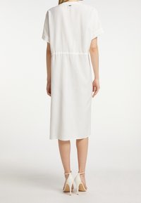Weißes Midikleid mit kurzen Ärmeln, tailliertem Schnitt und glatter Textur. Getragen mit weißen High-Heels. Sauberes, minimalistisches Design.