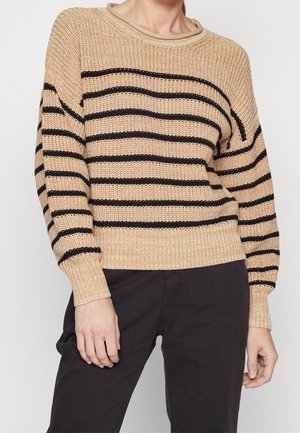 Beżowy sweter dziergany z czarnymi poziomymi paskami, noszony z czarnymi spodniami, widoczny od szyi do bioder, ręce swobodnie opuszczone wzdłuż ciała.