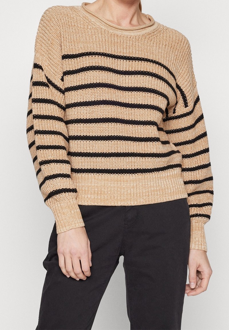 Pull en tricot beige avec des rayures horizontales noires porté avec un pantalon noir, montré du cou jusqu'aux hanches, les mains détendues le long du corps.