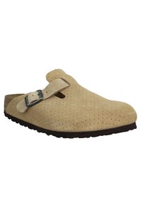 Sabot en suède beige avec un design perforé, une sangle à boucle et une semelle en caoutchouc marron. Il présente un bout rond et une texture lisse.